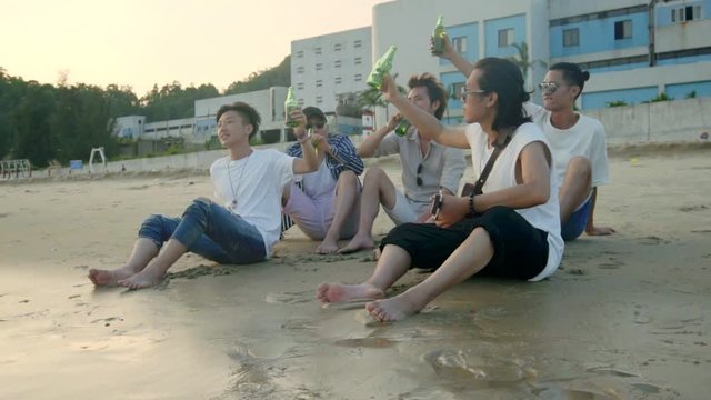 young asian adults sitting relaxing on beach and drinking beer to a toast, slow motion