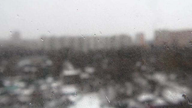 Water droplets on dirty glass.  Rain with snow. Rain drops strike a window pane and slowly flow down during a spring rain. Close up. 4K. Depressing picture of rain and grey blurred city in the back.