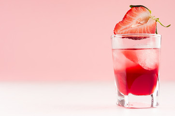 Exquisite cold red alcoholic liquor with ice cubes and strawberry slice in shot glass closeup on white wood table and pastel pink wall, copy space.