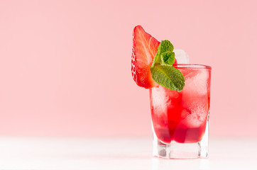 Summer fresh red cocktail in shot glass with ice cubes, strawberry slice, green mint on white wood table and pastel pink wall, copy space.