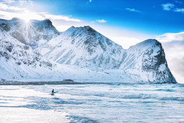 Iconic arctic surf beach - UNSTAD, located over polar circle in Norway, Moskenes on Lofoten islands. Man surfer going with surf board into Norwegian sea waters at epic rocks background. Winter season.