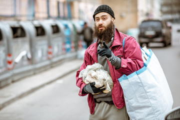 Portrait of a homeless bearded beggar standing with bag and some finded food near the trash...