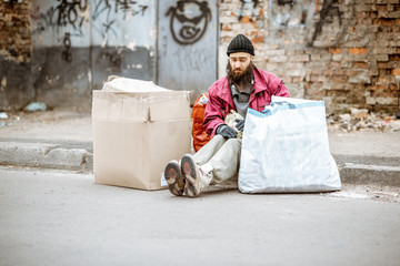 Homeless depressed beggar sitting with bags and cardboards on the street on the ragged wall...