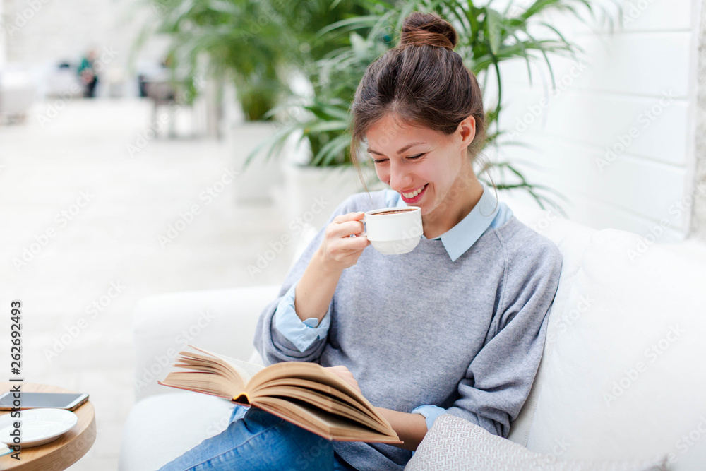 Wall mural Young woman is drinking coffee and reading book. Girl is smiling, enjoying relaxation and sitting on sofa in cozy modern cafe indoors.