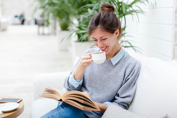 Young woman is drinking coffee and reading book. Girl is smiling, enjoying relaxation and sitting...