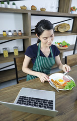 Beautiful young Asian woman wears apron eats breakfast on a wooden table in the dining room with laptop.