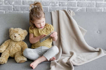 Little charming girl sits on a gray sofa. She are playing with tablet. Toy teddy is sitting next to her. The beige  plaid is lying on the back of the sofa. Behind the sofa we see a brick gray wall.