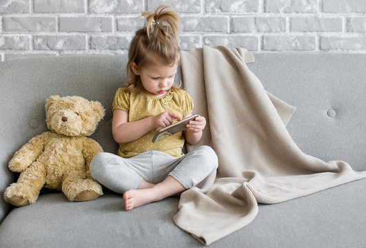 Little charming girl sits on a gray sofa. She are playing with tablet. Toy teddy is sitting next to her. The beige  plaid is lying on the back of the sofa. Behind the sofa we see a brick gray wall.