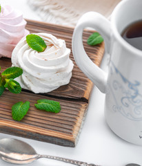 Handmade cream and strawberry marshmallows. Adorned with mint leaves. Tea in a white cup with a blue ornament. On a wooden background. Close-up.