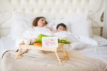 Obraz na płótnie Canvas Young mother with her little son relaxing in the bed. Mother's Day concept. Greeting card and breakfast in bed