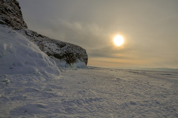 Rock on the shore of a snow-covered lake in the rays of sunset