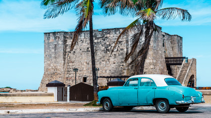 Havana, Cuba. Under the palm tree an old American car parked at it`s shade, besides of an old fort.