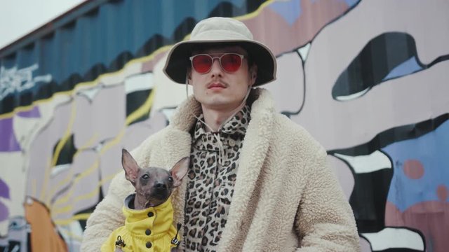 Portrait young man with hat in red sunglasses holding dog wear yellow clother in arms look at camera serious stylish fashion background artwall fun happy pet small young adult expression slow motion