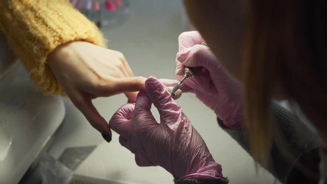The manicure expert wearing pink gloves removing shellac from the client nails with nail drill. Girl having black paited nails and tender hand. Only hands visible concept