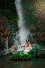 Sexy woman posing on the rock among green tropical plants and beside beautiful waterfall with blue water. Fairy tale.