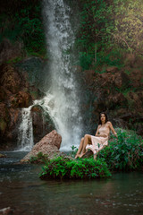 Sexy woman posing on the rock among green tropical plants and beside beautiful waterfall with blue water. Fairy tale.