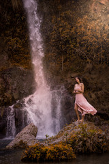 Fototapeta na wymiar Attractive girl posing on the rock among in fairy fast forest in autumn and beside beautiful waterfall with blue water. Fairy tale.
