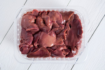 Raw chicken liver in a transparent container on a white wooden table. View from above.