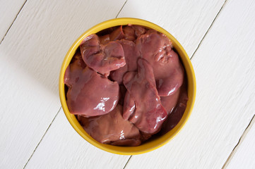 Raw chicken liver in a yellow bowl on a white wooden table. View from above.