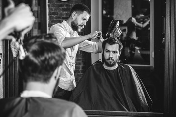 Young handsome barber making haircut for attractive bearded man at barbershop. Black and white or colorless photo. Hairstyle, salon, hairdresser, barber shop, lifestyle concept. Caucasian male models.