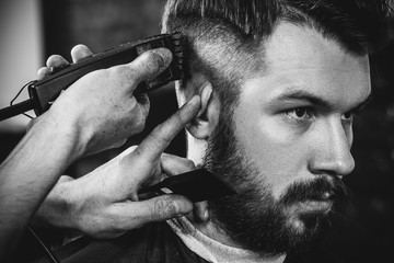 Young handsome barber making haircut for attractive bearded man at barbershop. Black and white or...