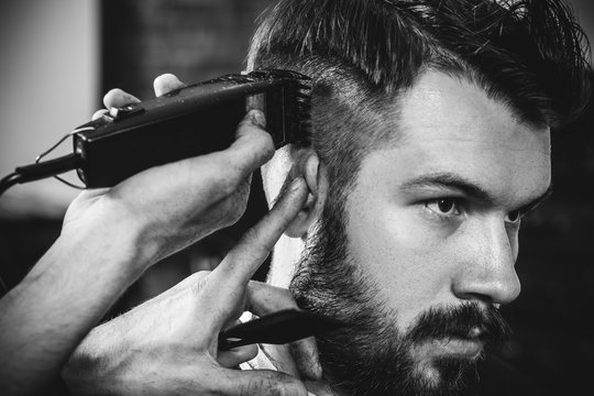 Young handsome barber making haircut for attractive bearded man at barbershop. Black and white or colorless photo. Hairstyle, salon, hairdresser, barber shop, lifestyle concept. Caucasian male models.