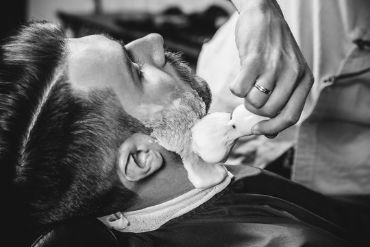 Client during beard shaving in barbershop. Young handsome barber and attractive bearded man. Black and white or colorless photo. Hairstyle, salon, hairdresser, barber shop, lifestyle concept