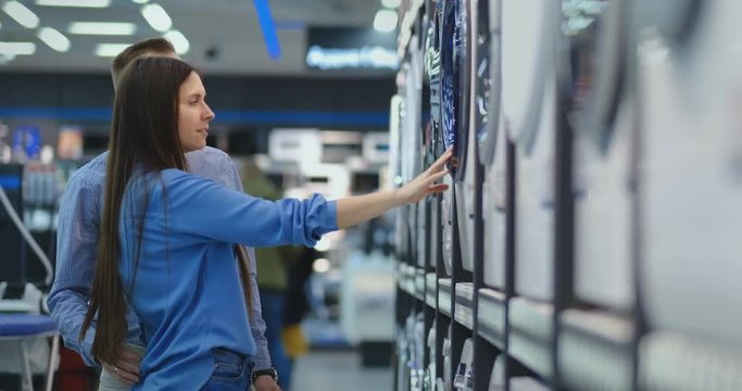 Beautiful married couple buys a new washing machine for their home. Open the door to the store to study the characteristics. Stand at the counter with electrical appliances