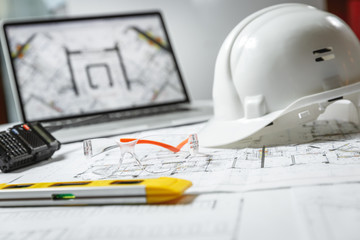 White hard hat, laptop  with drawings, glasses and walkie talkie with  and blueprints on a table