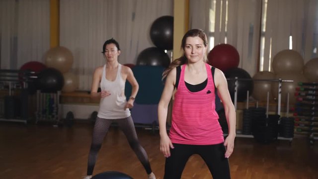 Two female athletes in a sport dress perform a step-up raising of their legs. Stretching and paletes are performed by a professional instructor in a personal lesson.