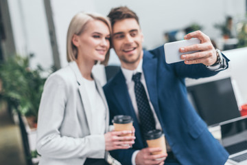 selective focus of two young business partners holding paper cups cups and taking selfie in office