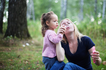 Mother and little daughter blowing soap bubbles in park. Happy and carefree childhood. Good family relations.