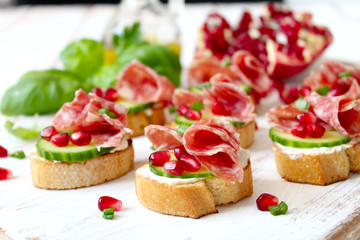 Fototapeta na wymiar Canape or crostini with toasted baguette, light cheese, cucumber, pomegranate and salami on light background. Top view with copy space.