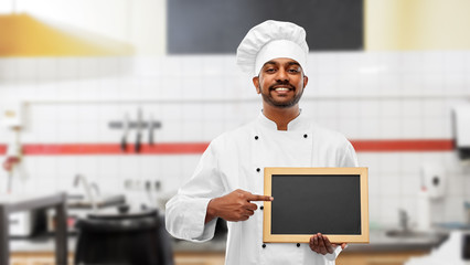 cooking, profession and people concept - happy male indian chef in toque with blank chalkboard for menu over restaurant kitchen background