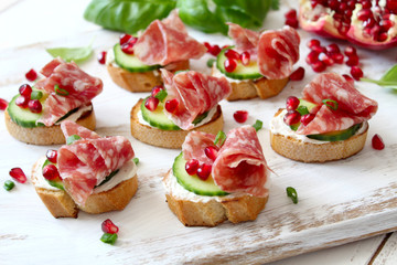 Canape or crostini with toasted baguette, light cheese, cucumber, pomegranate and salami on light background. Top view with copy space.