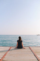 Girl sitting on the pier and looking at the sea horizon and blue sky. Vacation at sea. Summer time sea vacation background 