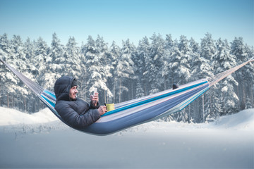 A smartphone user sits in a hammock in a winter snowy forest. Mug with a hot drink in his hand