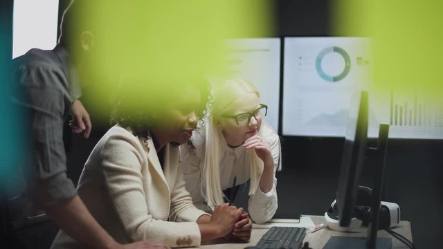 international teamwork in modern office, man and two women managers look at the screen with financial graphics in monitor, camera movement through glass with sticky notes.