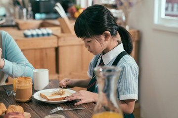 focus on smart cute little girl having breakfast wear uniform before school in morning. daughter sitting beside mom putting peanut jam on bread toast. family time healthy lifestyle early day.