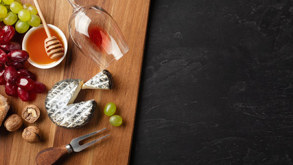 Cheese head, bunch of grapes, honey, nuts and wineglass on wooden board and black background