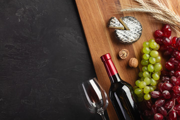 Red wine bottle, bunch of grapes, cheese, ears of wheat and wineglass on wooden board and black background