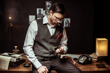 Concentrated detective in glasses sitting on wooden table and looking at pocket watch in office