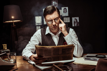 Concentrated detective in glasses reading newspaper in dark office
