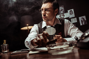 Pensive detective in glasses holding magnifier and looking away in office