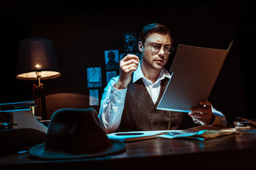 Concentrated detective in glasses reading dossier in dark office