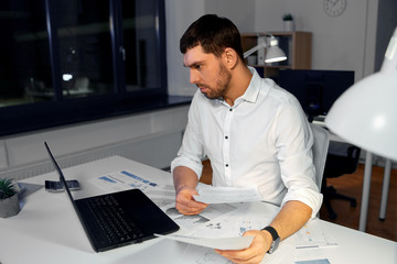 business, deadline and stress concept - businessman with papers and laptop computer working at night office