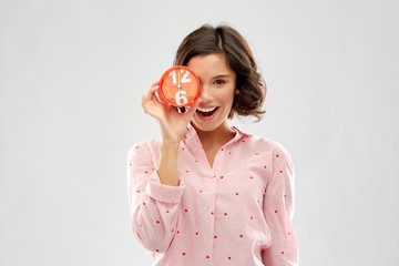 people and bedtime concept - happy young woman in pajama covering one eye with alarm clock showing six o'clock over grey background