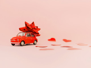 Red retro toy red car with red bow for Valentine's day on pink background with heart confetti. February 14 card. 8 March,International Women's Day. Selective focus