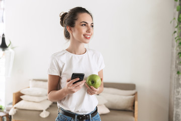 Happy beautiful young student girl eat apple posing indoors using mobile phone.