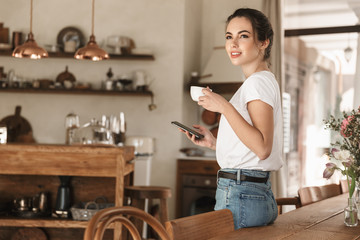 Beautiful young student girl drinking coffee posing indoors chatting by mobile phone.